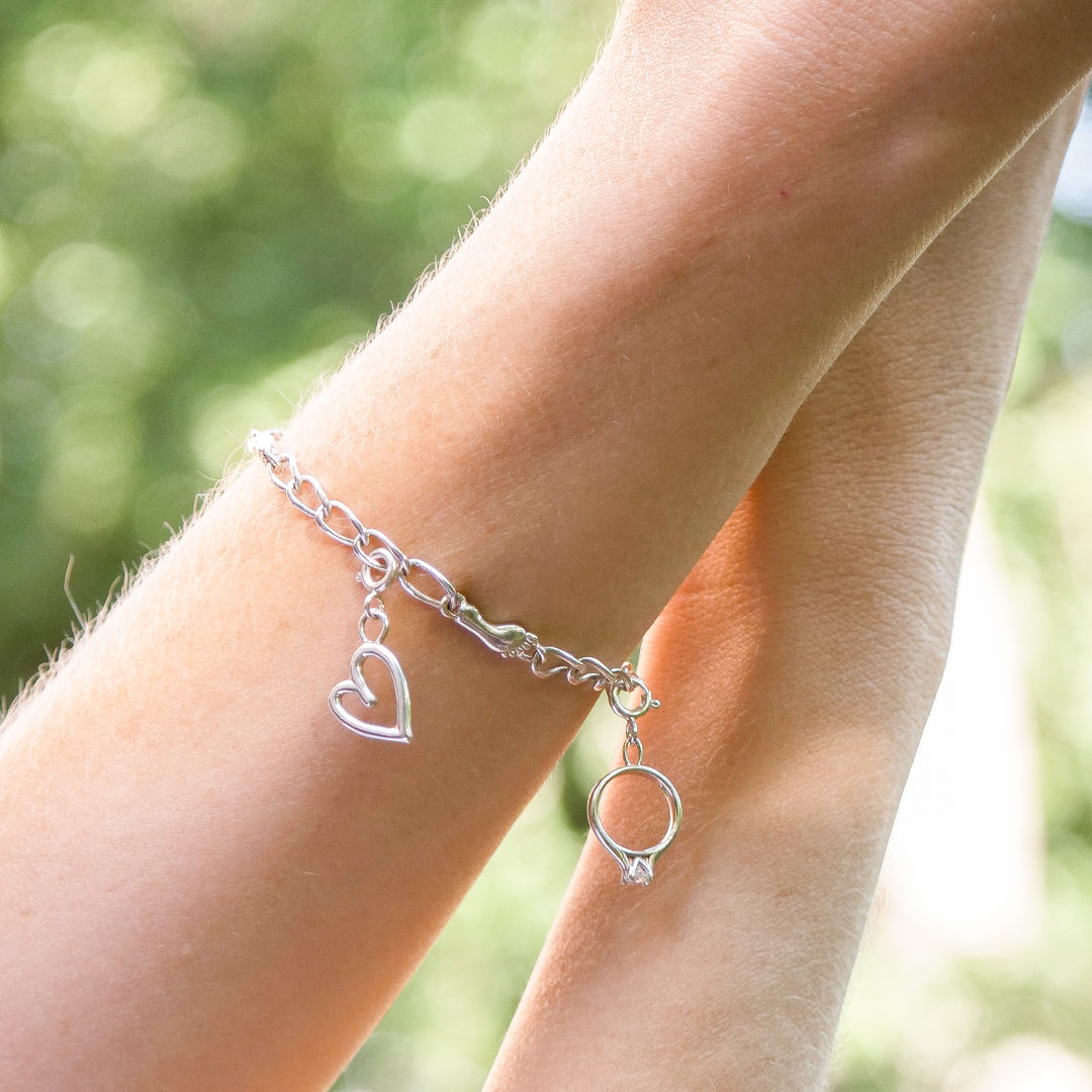 Silver charm bracelet with heart and ring charms worn on wrist against blurred natural background