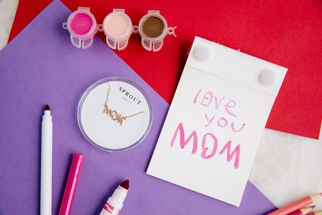 Rose gold handwriting necklace spelling “MOM” displayed with child’s card saying “Love you Mom.”
