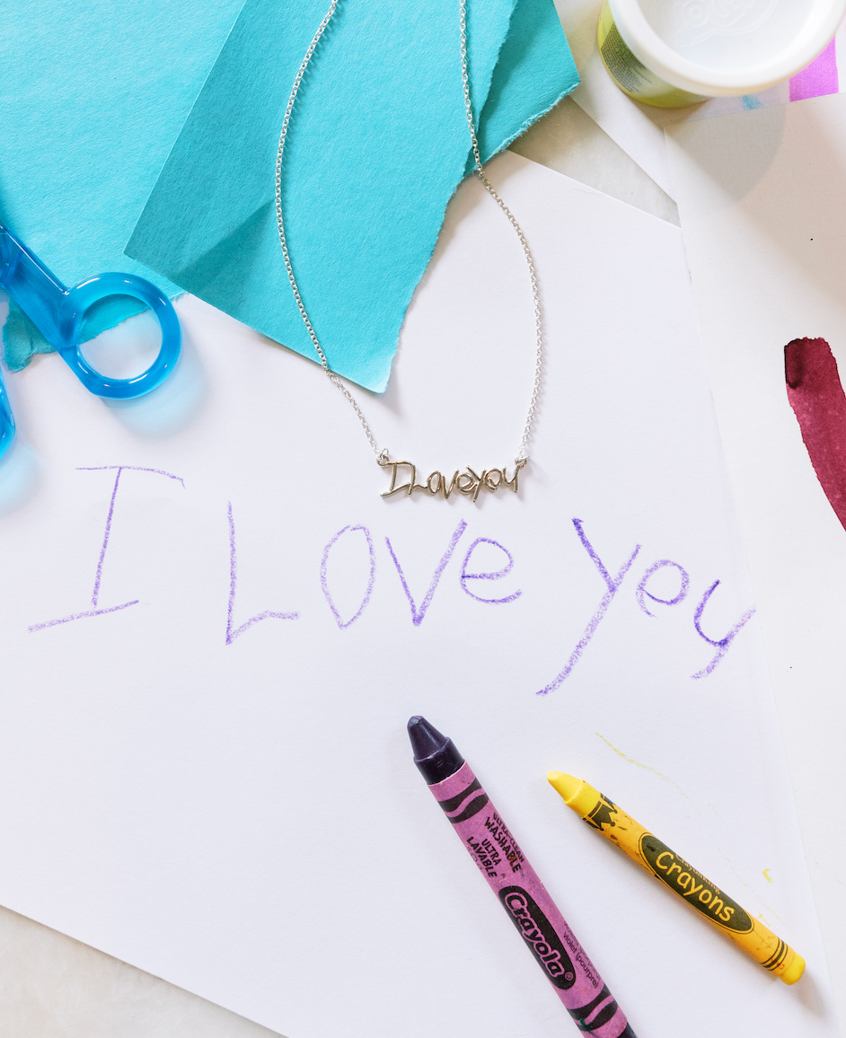 Custom handwriting necklace displayed with child’s drawing and crayons spelling “I love you.”